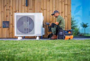 A technician is servicing an air conditioning unit located beside a modern home. Dressed in work attire, he uses tools from an organized toolbox while seated on the grass. The setting features a wooden wall and bright blue windows, indicating a well-maintained property.